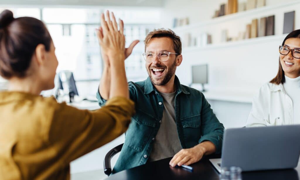 one man, two woman sitting in a round in the office, smiling and giving high five to each other